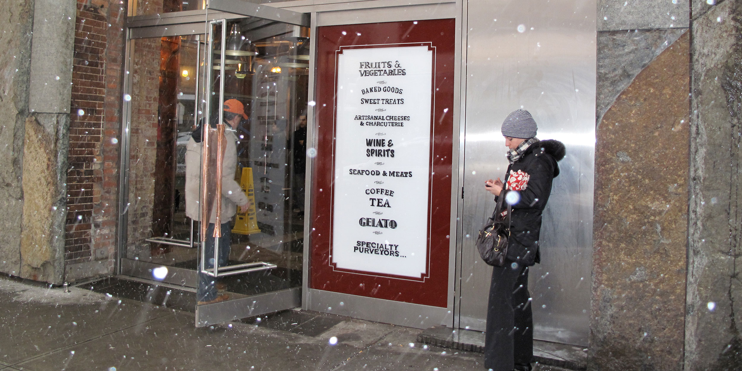 A person in dark clothing taking a photo of a storefront display with a promotional poster.