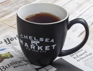 A black mug with "CHELSEA MARKET" text filled with coffee, sitting on a newspaper.