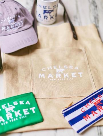 Market tote bag with purple cap, coffee mug, and market-branded items on marble surface.